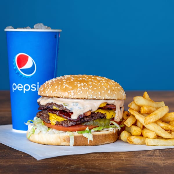 Double cheeseburger with bacon, fries, and a Pepsi drink on a wooden table.