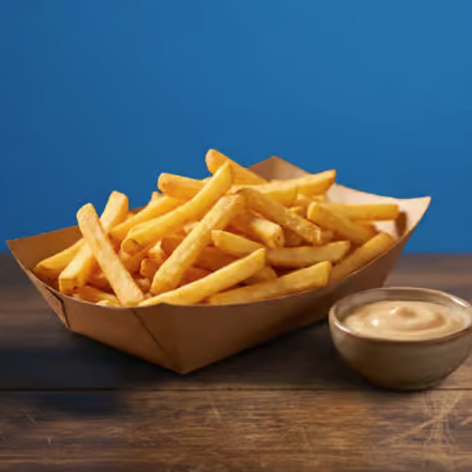 Basket of golden French fries with dipping sauce on a rustic wooden table against a blue background.